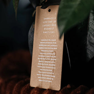 Loodusliku välimusega pruun pabersilt või etikett valge tekstiga. Sildil on inspireeriv sõnum: "EMBRACE A LIFETIME OF LUXURY AND WARMEST EMOTIONS". Järgneb pikem tekst, mis kirjeldab toote looduslikku päritolu ja traditsioone, mainides alpaka ja villa kasutamist luksuslike rõivaesemete valmistamisel. Tekst viitab Andide pärandile ja Põhjamaade sensibiliteedi kombineerimisele, et pakkuda ajatut luksust ja vastupidavat stiili. Sildil on ülaosas auk, et seda saaks kinnitada toote külge. | Trükikoda Brandcafe