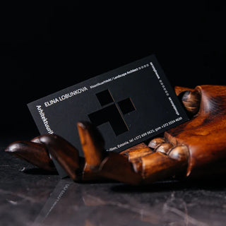 Black business card for landscape architect Elina Lobunkova with a die-cut plus-shaped cutout and white printed text, held by a wooden artist's mannequin hand on a dark marble surface.