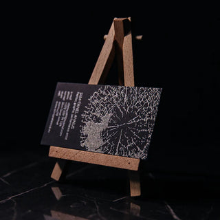 Black business card with a silver foil tree-ring pattern and an overlaid map silhouette, displayed upright on a small wooden easel against a dark marble surface.