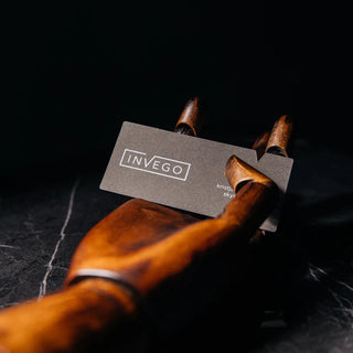  Gray business card with a white "INVEGO" boxed logo and name text, held between the fingers of a wooden artist's mannequin hand on a dark marble surface.