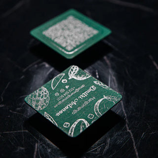 Two green square business cards with silver foil tropical fruit illustrations and text on one card and a QR code on the other, placed on a dark marble surface.