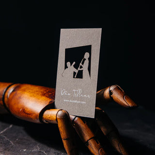 Kraft-colored business card with a laser-cut window revealing silhouettes of two musicians playing, held by a wooden artist's mannequin hand, with "Duo Telluur" script and website text below.