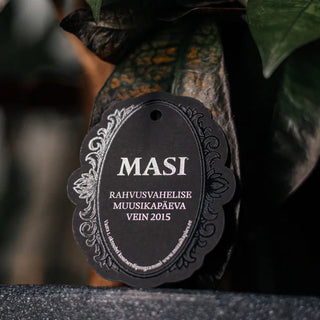 Elegantne tume silt või etikett kaunistatud ornamentidega äärisel. Sildil on kirjas "MASI" ja "RAHVUSVAHELISE MUUSIKAPÄEVA VEIN 2015". Tõenäoliselt on tegemist piiratud koguses veinipartii või erisildiga, mis on loodud rahvusvahelise muusikapäeva tähistamiseks 2015. aastal. Musta tausta ja hõbedase või valge trükiga kujundus loob piduliku ja luksusliku mulje. Sildil on ülaosas auk, et seda saaks kinnitada pudeli või pakendi külge. | Trükikoda Brandcafe