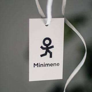 Valge etikett või silt mustade elementidega. Keskel on stiliseeritud inimfiguuri kujutav logo või sümbol, mille all on tekst "Minimene". Sildil on ülaosas auk valge nööri või paelaga, et seda saaks kinnitada toote külge. Lihtne ja minimalistlik disain, mis võib olla seotud lasteriietega, mänguasjadega või muude lastele suunatud toodetega, millele viitab mängulise inimfiguuri kujutis ja nimi. | Trükikoda Brandcafe