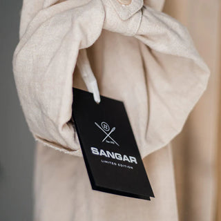 Elegantne must toote silt tekstiga "SANGAR LIMITED EDITION". Sildil on Sangari brändi logo koos õmblusvahendite sümboolikaga (nõel ja niit). Riideeseme külge kinnitatud silt, mis rõhutab piiratud koguses kollektsiooni. Must taust ja valge või hõbedane trükk loovad luksusliku mulje. Pildil on näha osa heledast riideesemest, millele silt on kinnitatud. | Trükikoda Brandcafe