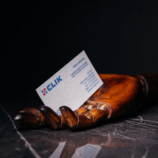 White business card with a blue "CLIK" logo and red icon, printed contact details in blue text, resting between the fingers of a wooden artist's mannequin hand on a dark marble surface.